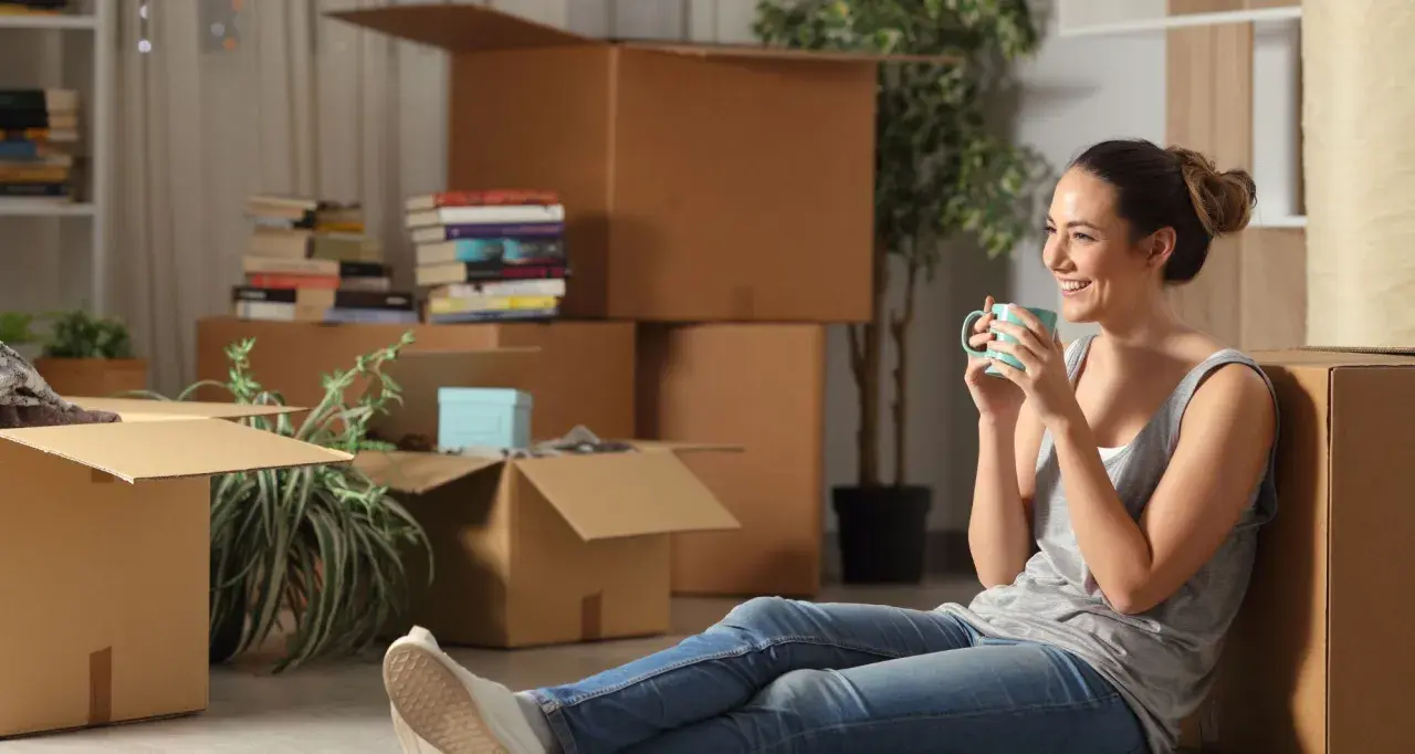 Une femme boit une tasse de café, entourée de nombreux cartons de déménagement.