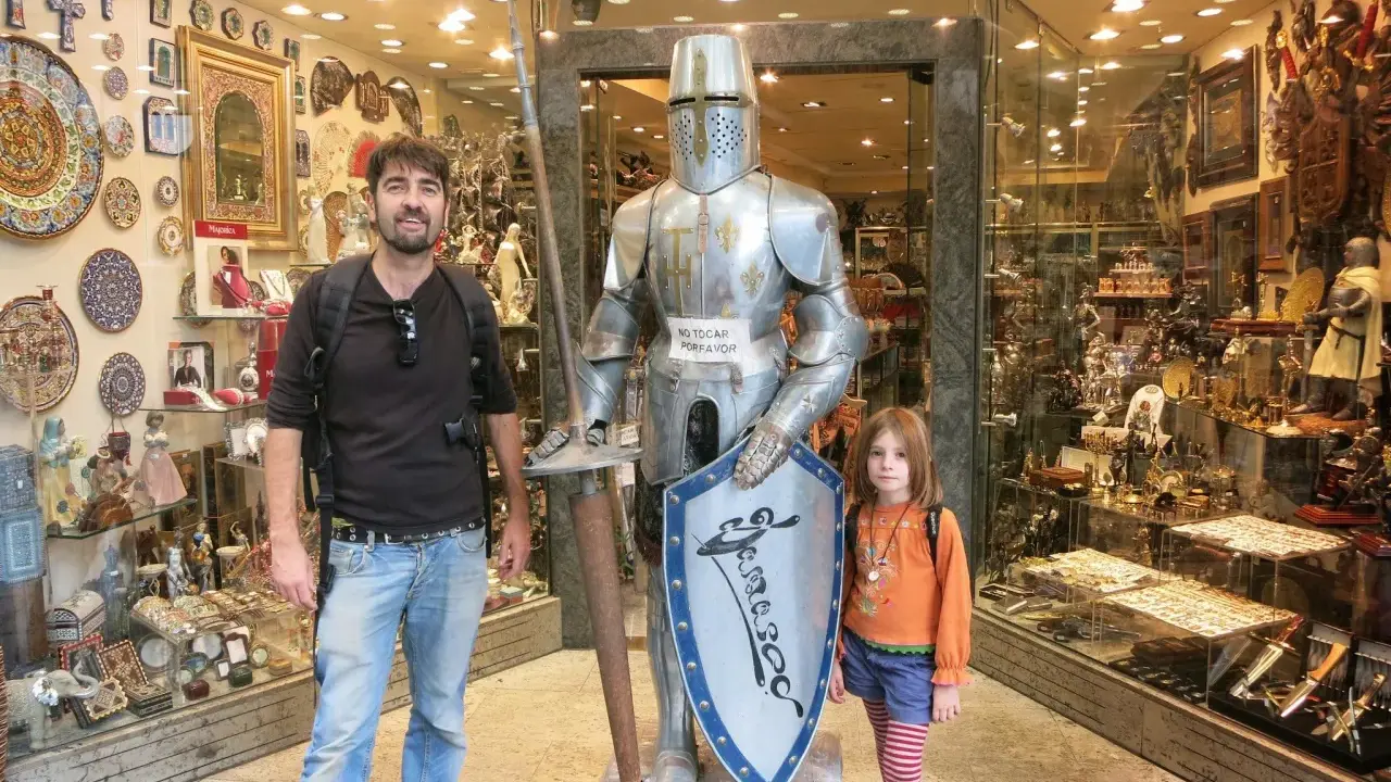Un padre y su hija posan junto a una armadura de caballero en una tienda de souvenirs en Toledo con niños.