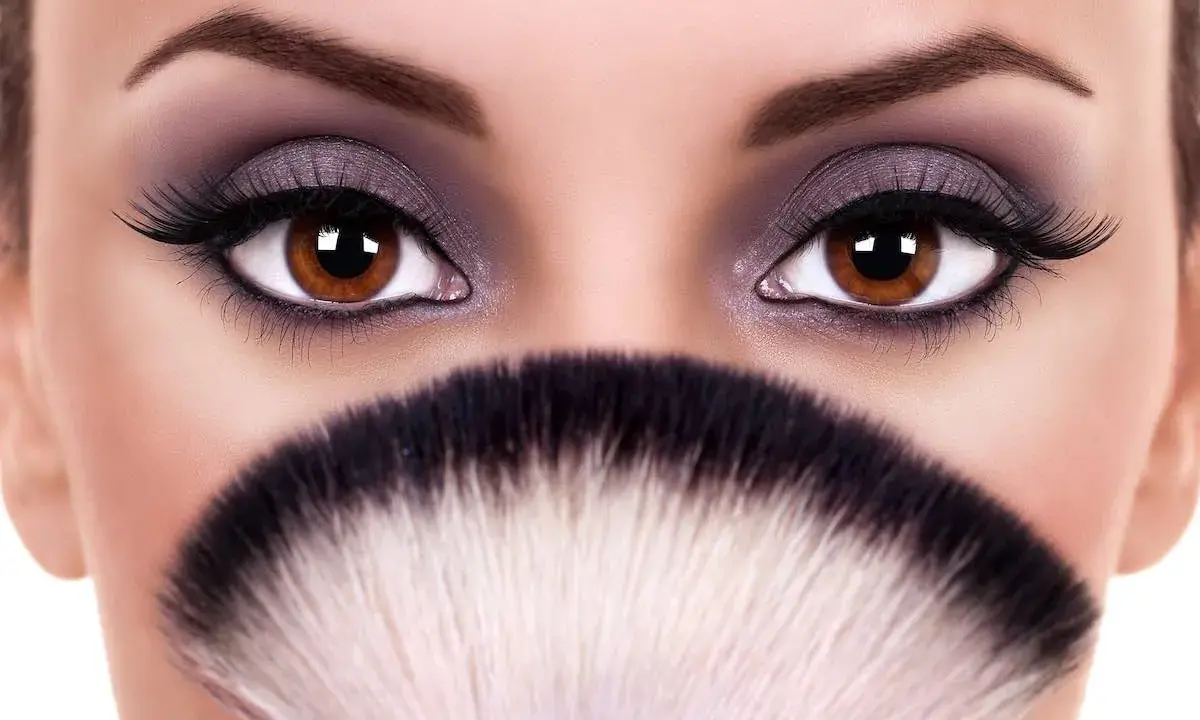 Occhi grandi e tondi esaltati da un trucco smokey viola e ciglia finte, con un pennello da trucco in primo piano.