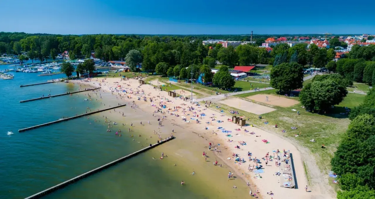Słoneczna plaża w Giżycku tętni życiem. Ludzie odpoczywają na piasku, kąpią się w jeziorze i spacerują wzdłuż drewnianych pomostów.