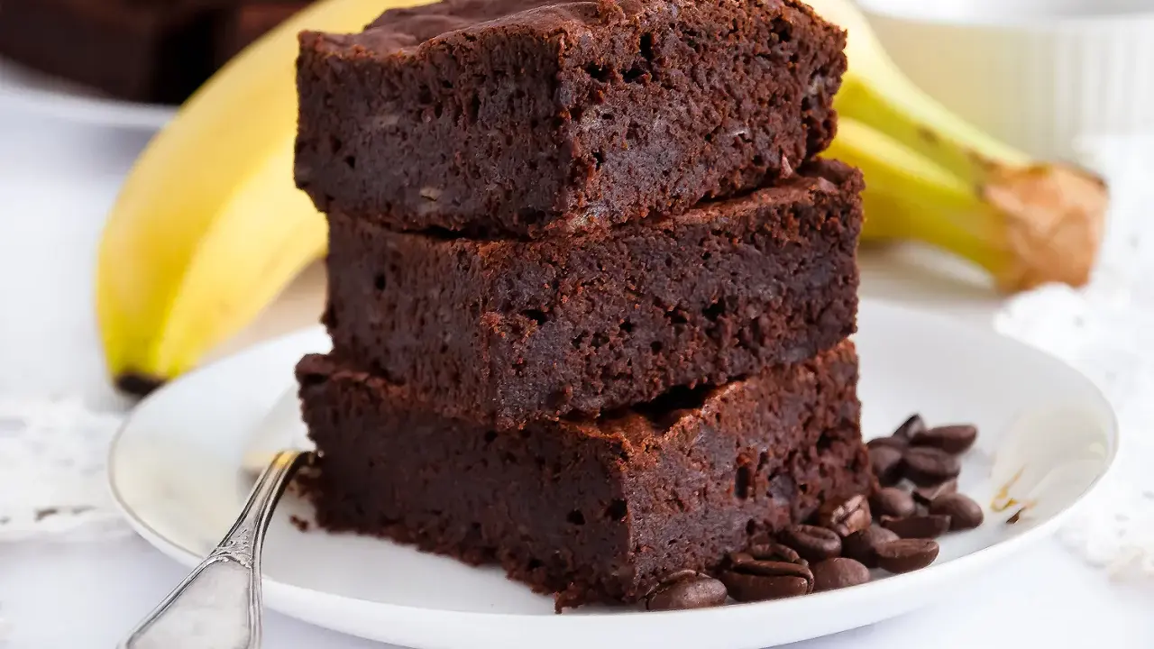 Stos puszystych brownie z bananami, ozdobiony ziarnami kawy, na talerzu z bananem w tle.
