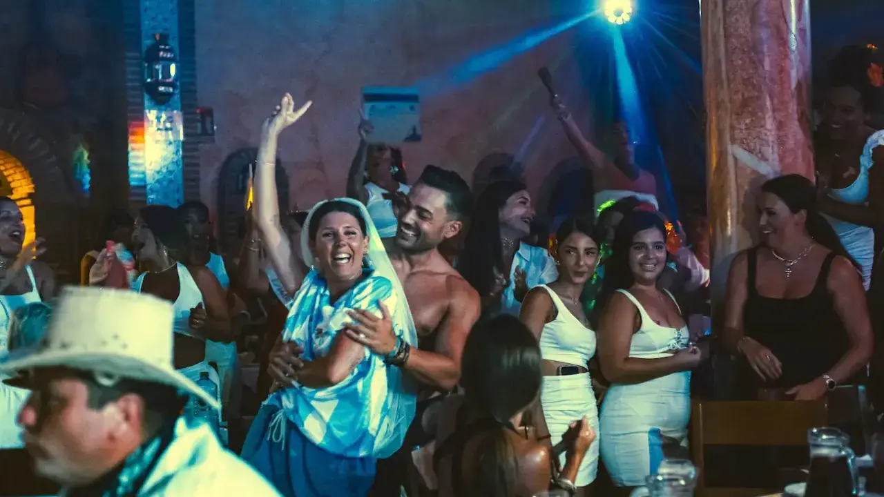 Gente celebrando y bailando en una fiesta, con luces de discoteca y un ambiente festivo. Parece una despedida de soltero con juegos y mucha alegría.