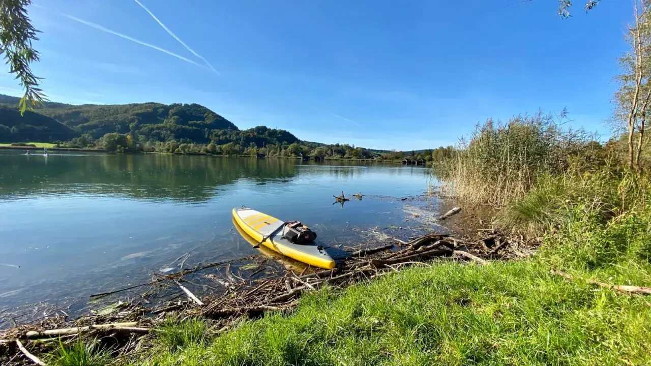 Gelbes SUP-Board liegt am Ufer bereit für SUP Touren Bayern. Ruhiger See, bewaldete Hügel und blauer Himmel.