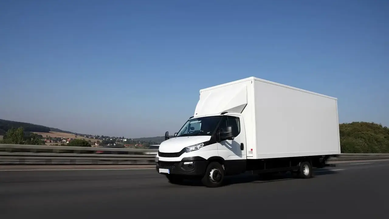 Biały samochód ciężarowy powyżej 3,5 tony pędzi autostradą. W tle krajobraz z pagórkami i domami.