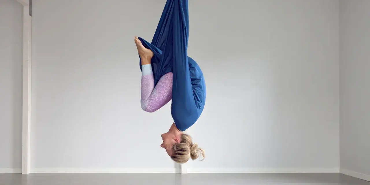 Kvinna hänger upp och ner i en blå tyghängmatta, utövar flying yoga.