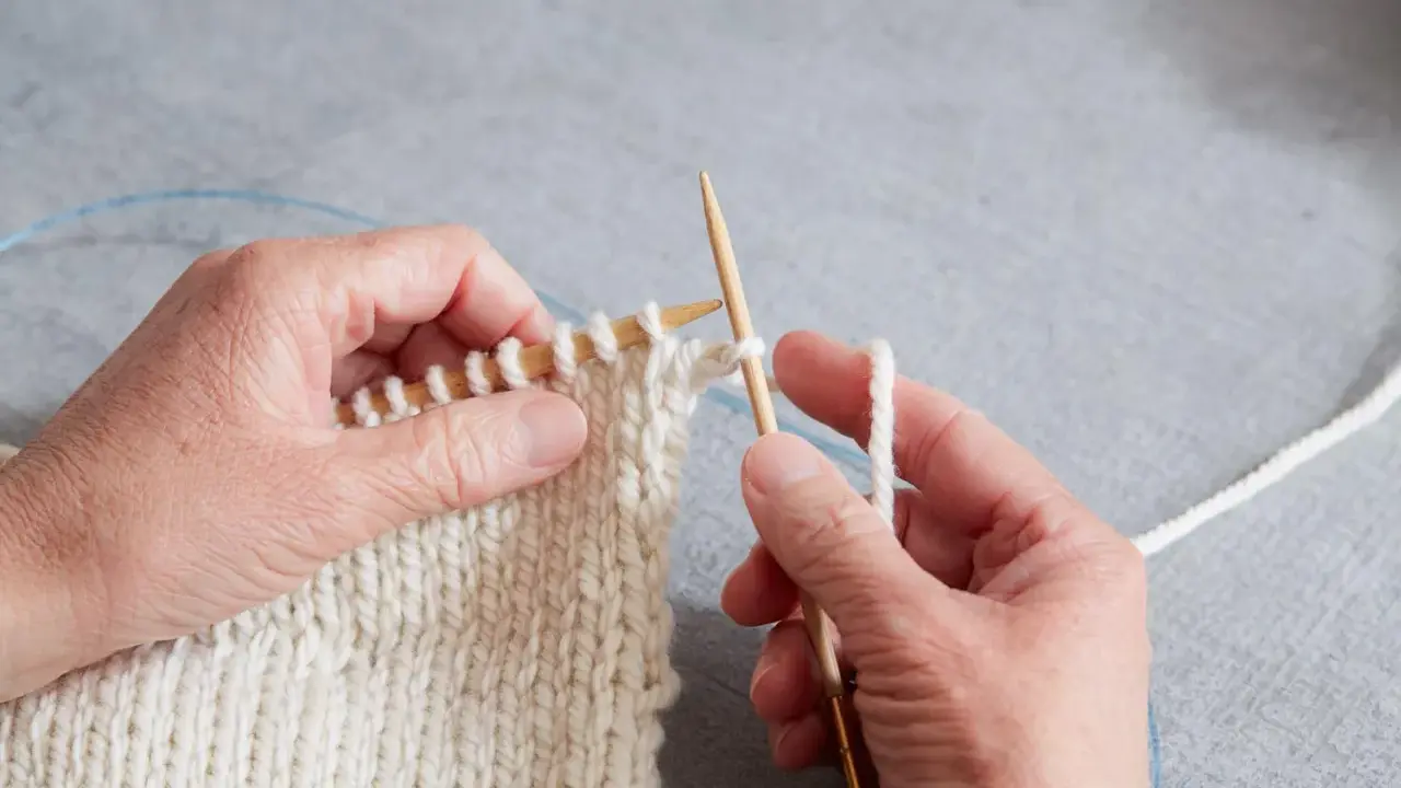 Händer stickar en vit resår med trästickor. En blå kabel syns i bakgrunden.