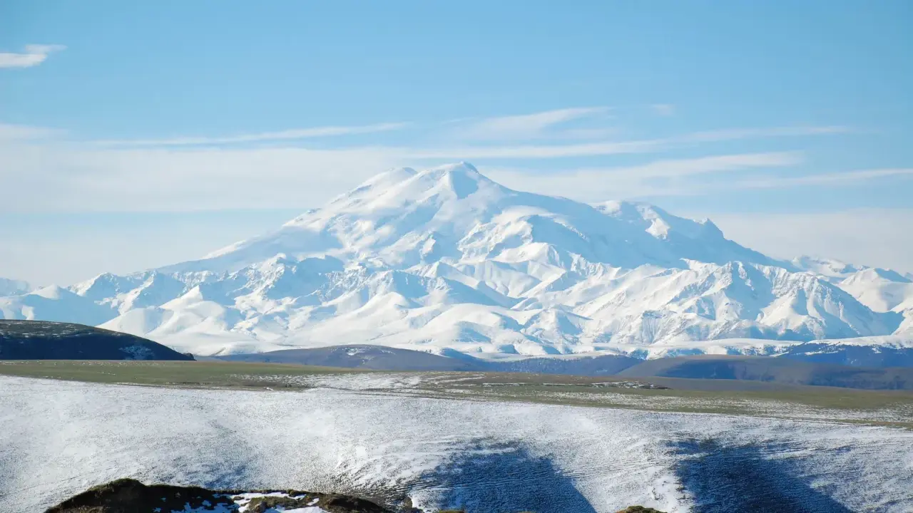 Pokryty śniegiem najwyższy masyw Wielkiego Kaukazu, Elbrus, góruje nad ośnieżonymi wzgórzami i zielonymi dolinami.
