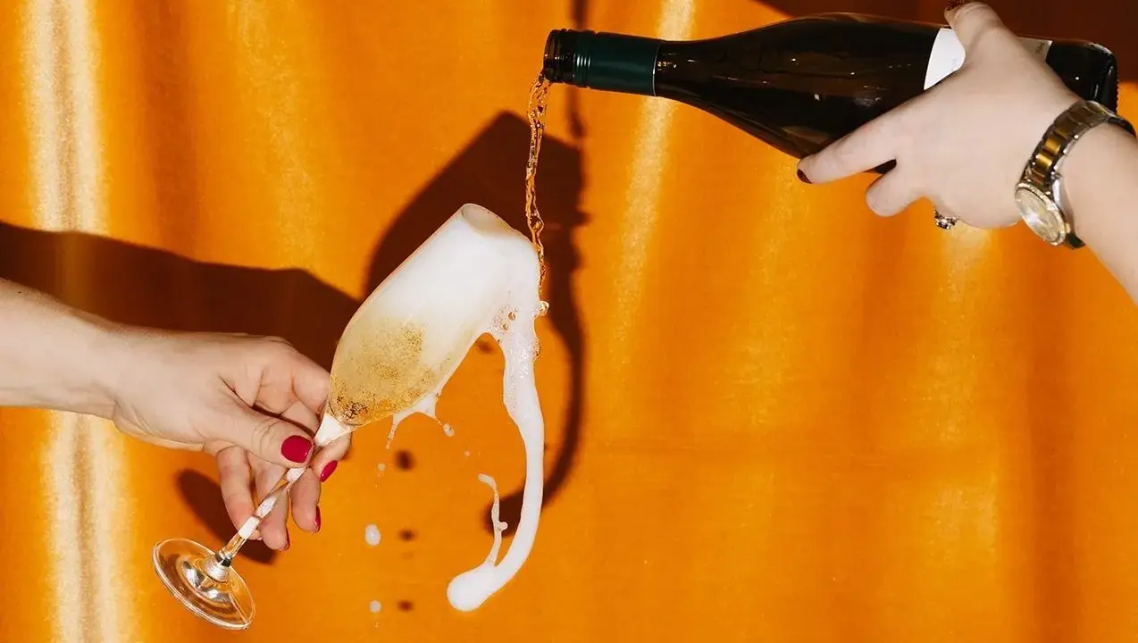 Schaumiges Sektglas läuft über, während eine Flasche auf orangefarbenem Hintergrund eingegossen wird. Eine Szene, die an die **zero dosage definition** erinnert, wo jeder Tropfen zählt.