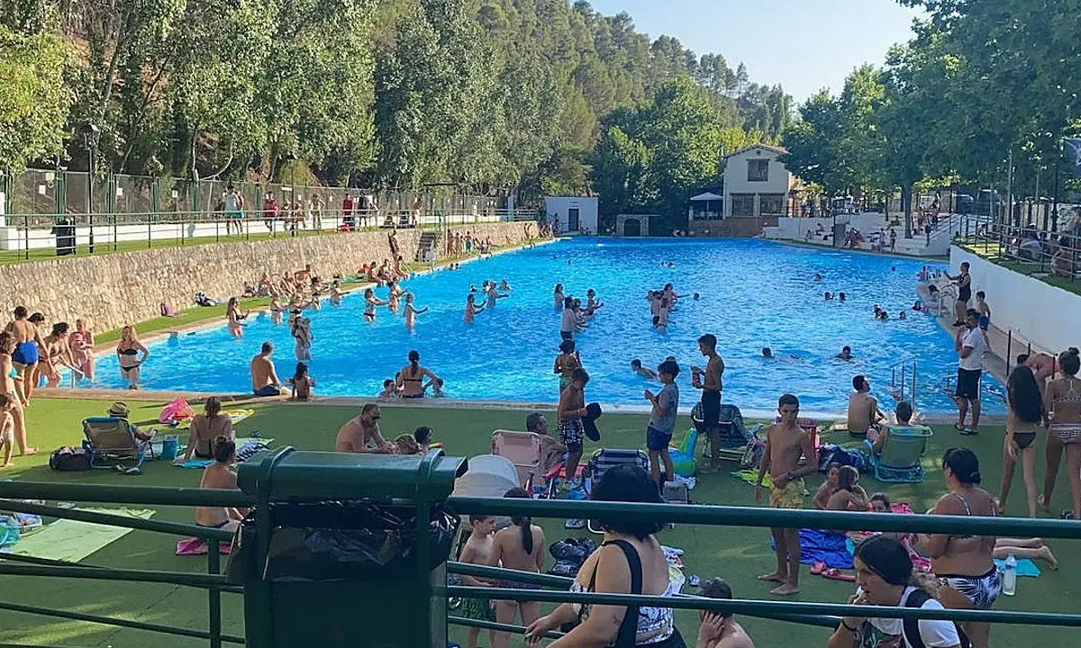 Gente disfrutando de un día soleado en la **piscina segura de la sierra**. Niños y adultos se refrescan en el agua azul, rodeados de árboles y un paisaje montañoso.