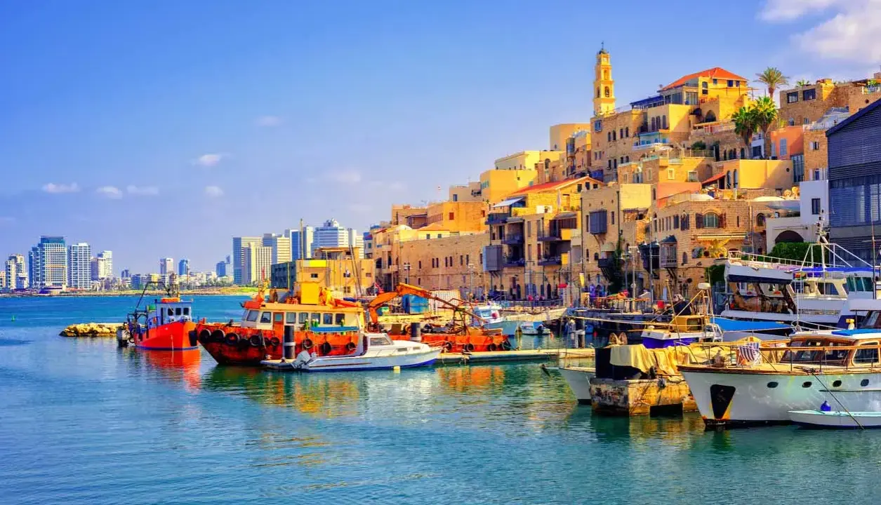 Bunte Boote im Hafen von Tel Aviv, mit der historischen Altstadt im Hintergrund und modernen Hochhäusern am Horizont.
