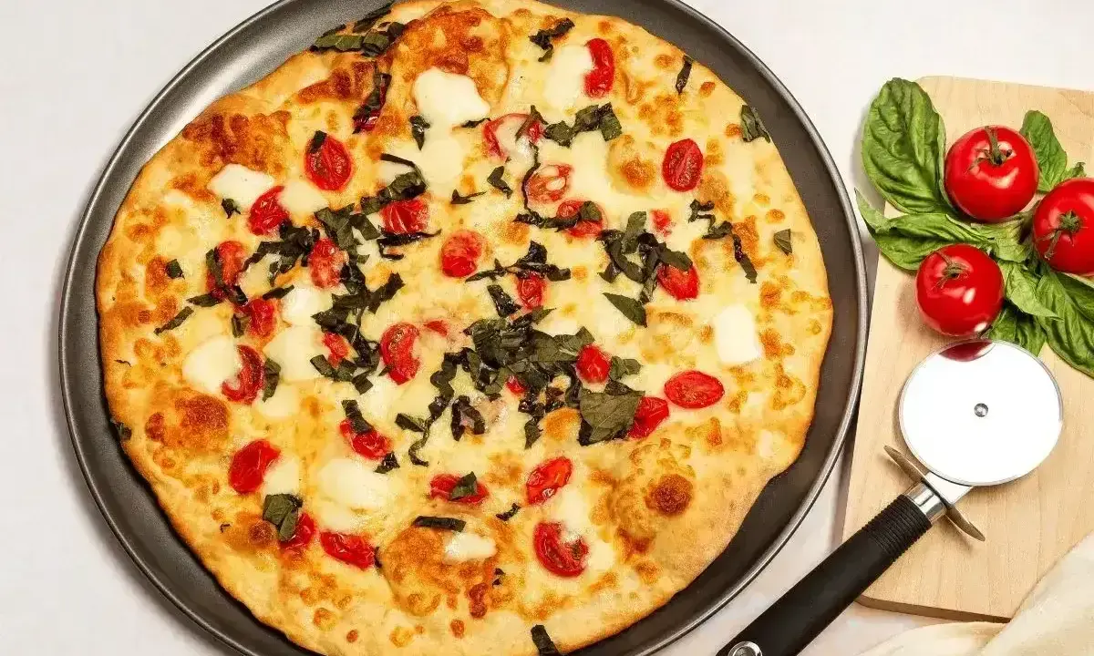 Pyszna pizza z serka wiejskiego, pomidorkami koktajlowymi i bazylią. Obok leżą świeże pomidory i krajalnica do pizzy.