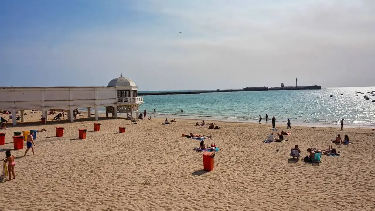 Sonniger Cadiz Strand mit Menschen, die sich am Meer entspannen. Ein weißes Gebäude mit Kuppel steht am Ufer.