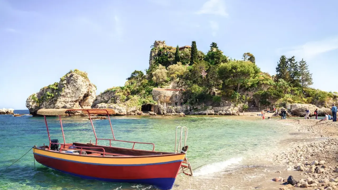 Ein Boot liegt im kristallklaren Wasser vor einer felsigen Insel mit üppiger Vegetation. Das sizilien wetter im oktober ist perfekt für einen entspannten Tag am Strand.
