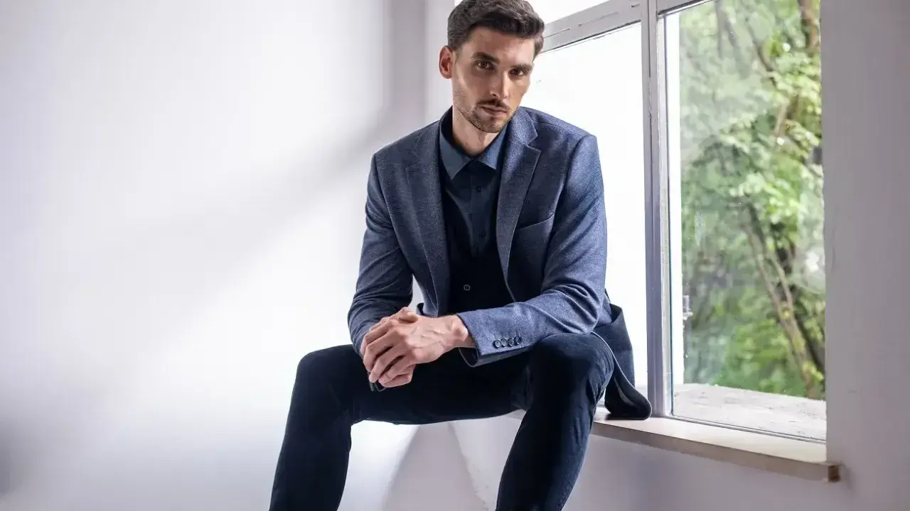 Elegancki mężczyzna w granatowej koszuli i marynarce, zastanawia się, jakie spodnie do niebieskiej koszuli będą najlepsze.