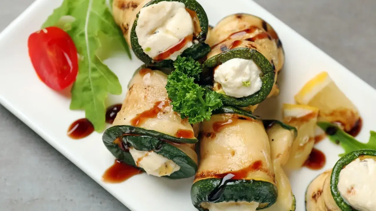 Deliziosi involtini di zucchine grigliate ripieni di formaggio cremoso, guarniti con pomodorini, rucola e salsa scura.