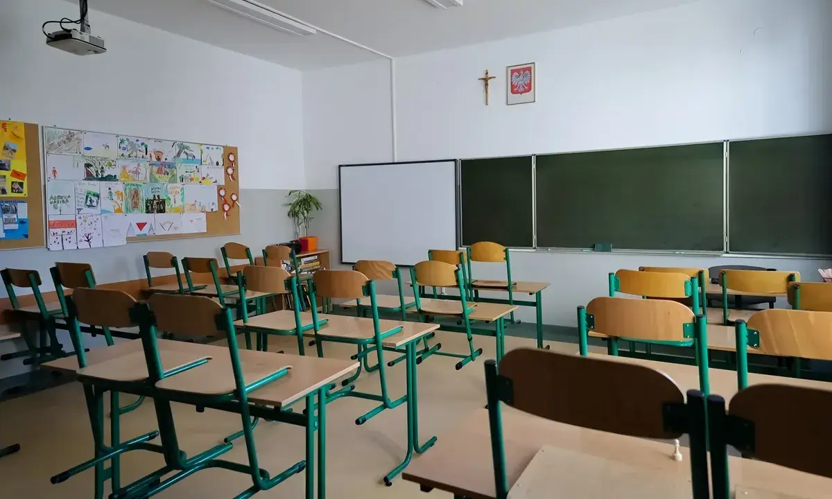 Pusta klasa z rzędami ławek i krzeseł, tablicą i dziecięcymi rysunkami na ścianie. Brak ucznia w tej sali może sugerować nieklasyfikowanie ucznia.