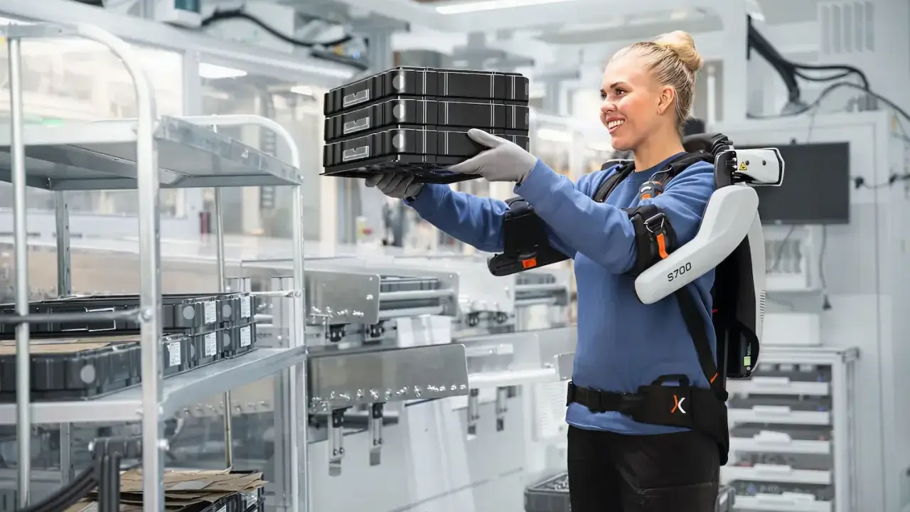 Une femme souriante porte des boîtes, aidée par un exosquelette, illustrant la définition de l'exosquelette comme aide à la manutention.
