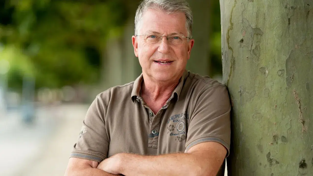 Carsten Sostmeier, ein Mann mit Brille und grauem Haar, lehnt lächelnd an einem Baum.