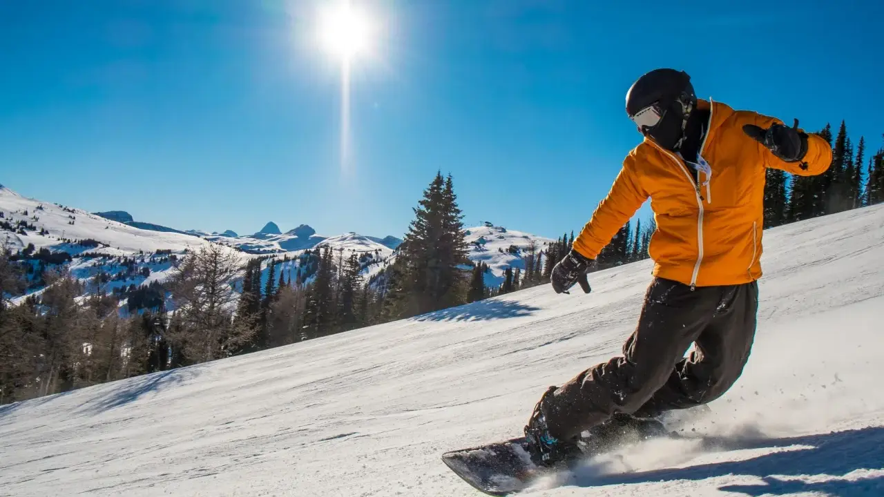 Snowboardzista w pomarańczowej kurtce zjeżdża po stoku, unosząc śnieg. To świetny przykład, jak jeździć na snowboardzie.
