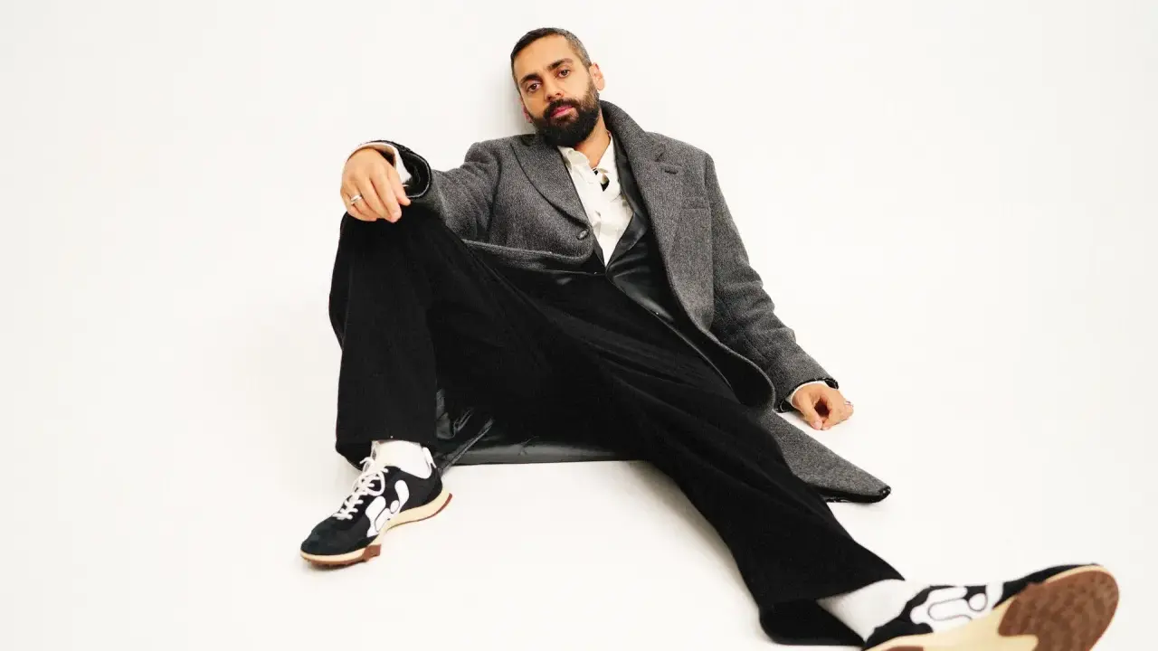 Ardalan Esmaili sitter avslappnat i en grå rock, vit skjorta och svarta byxor. Hans sneakers har en unik design.