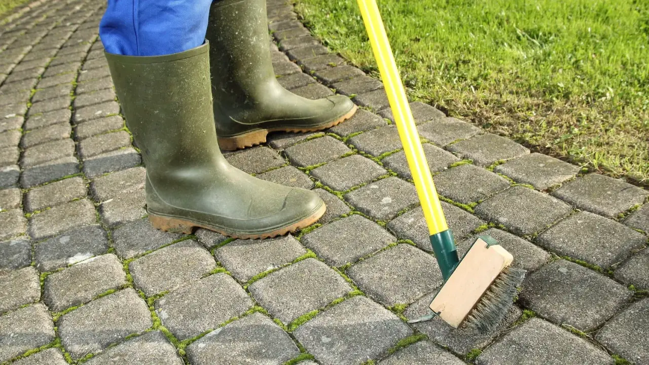 Person en bottes vertes brossant la mousse sur une terrasse pavée. Astuce pour savoir comment enlever la mousse sur une terrasse.