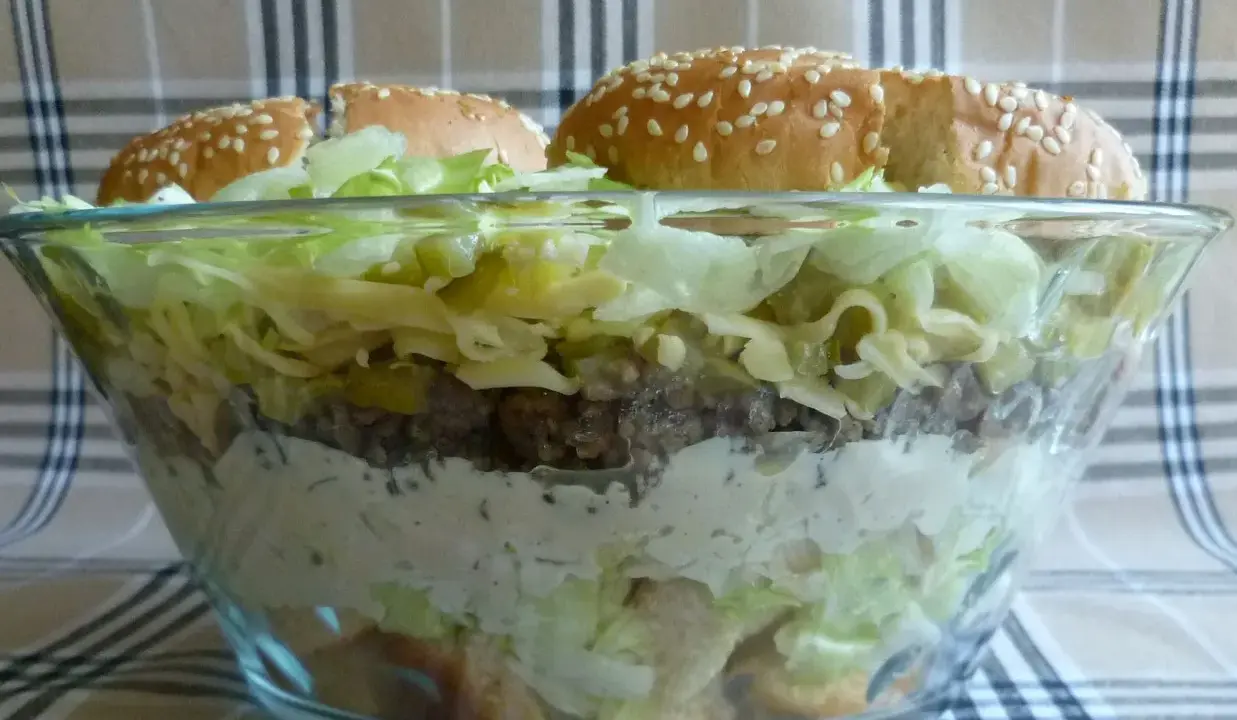 Warstwowa sałatka big mac w szklanej misie, z bułkami na wierzchu.