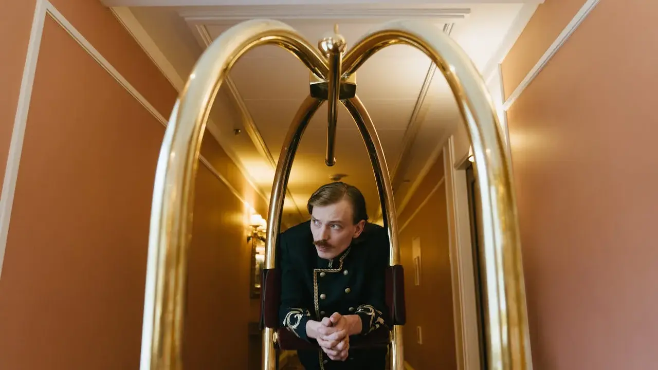 Elegancki hotelarz w historycznym mundurze czeka przy wózku bagażowym w hotelowym korytarzu.