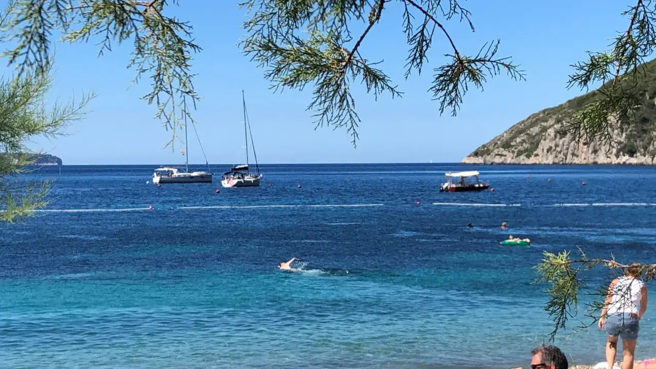 En man sitter på en stenstrand vid havet. I vattnet simmar en person och flera båtar ligger för ankar. En grön kulle reser sig i bakgrunden. En idyllisk vy från Vis, Kroatien.