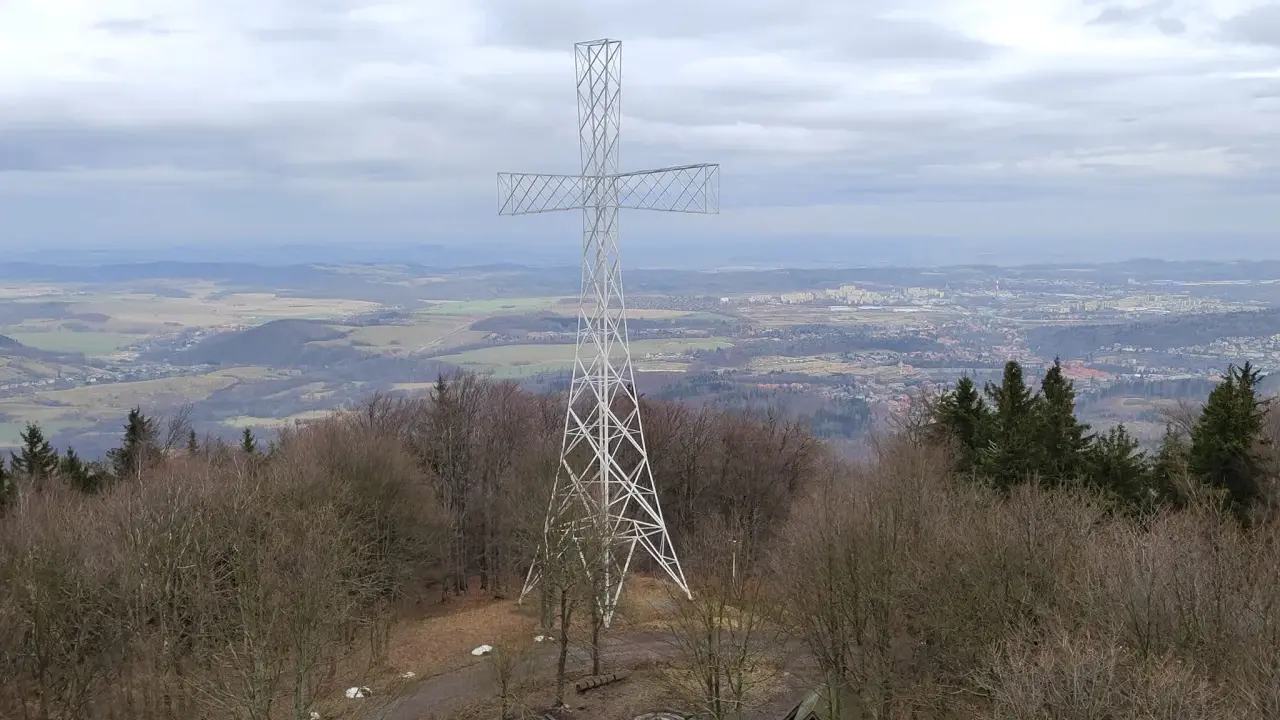 Wielki metalowy krzyż góruje nad lasem na Chełmcu. W dole rozciąga się panorama miasta i pagórkowatego krajobrazu.