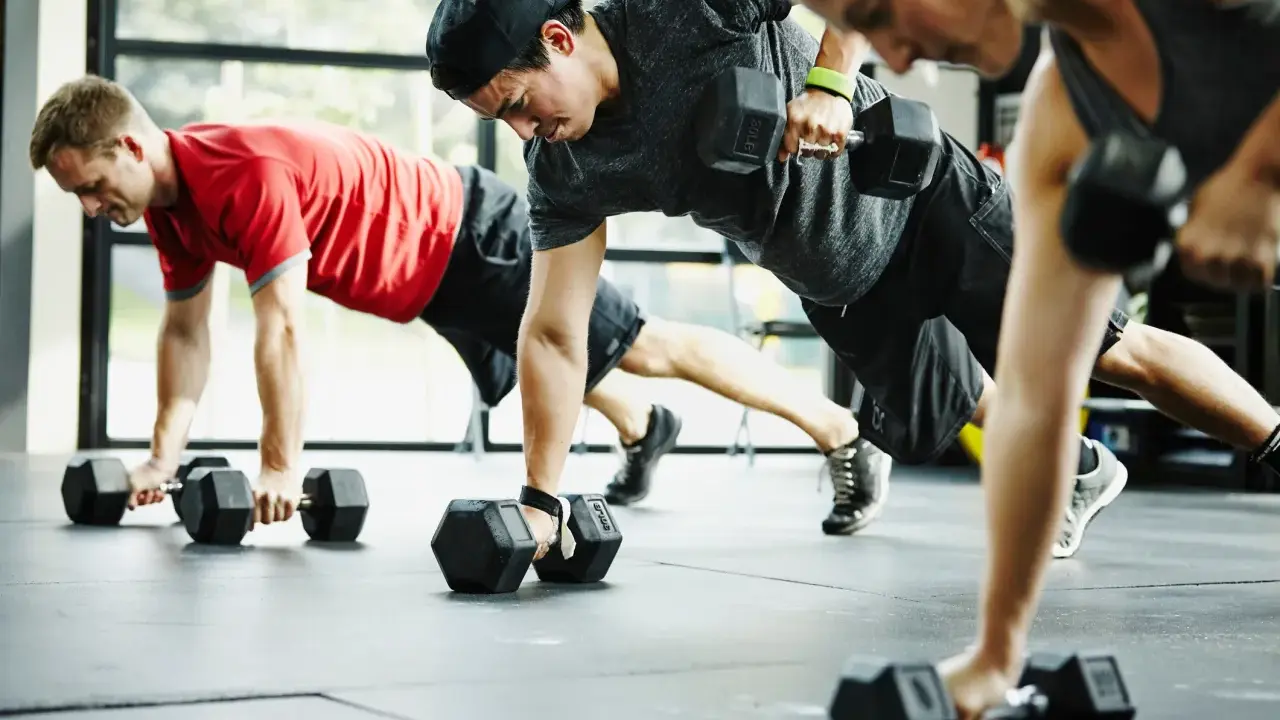 Drei Personen machen eine Plank-Übung mit Kurzhanteln. Ein Mann in Rot, ein Mann mit Kappe und eine Frau in Grau trainieren. CrossFit gesund und stark.