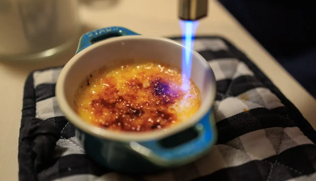 Flamme bleue caramélisant le sucre d'une crème brûlée, rappelant son origine.