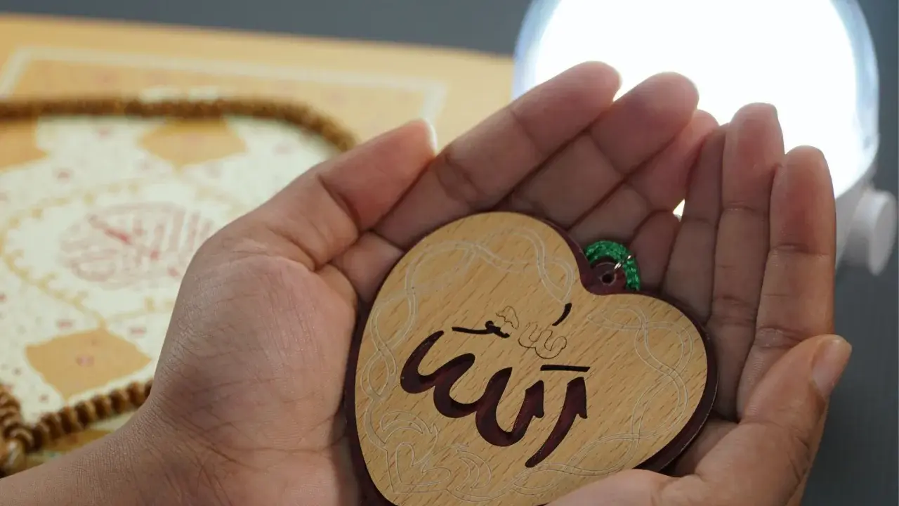 Hände halten ein Herz mit dem Schriftzug "Allah" in arabischer Schrift. Im Hintergrund liegen Gebetskette und ein Buch.