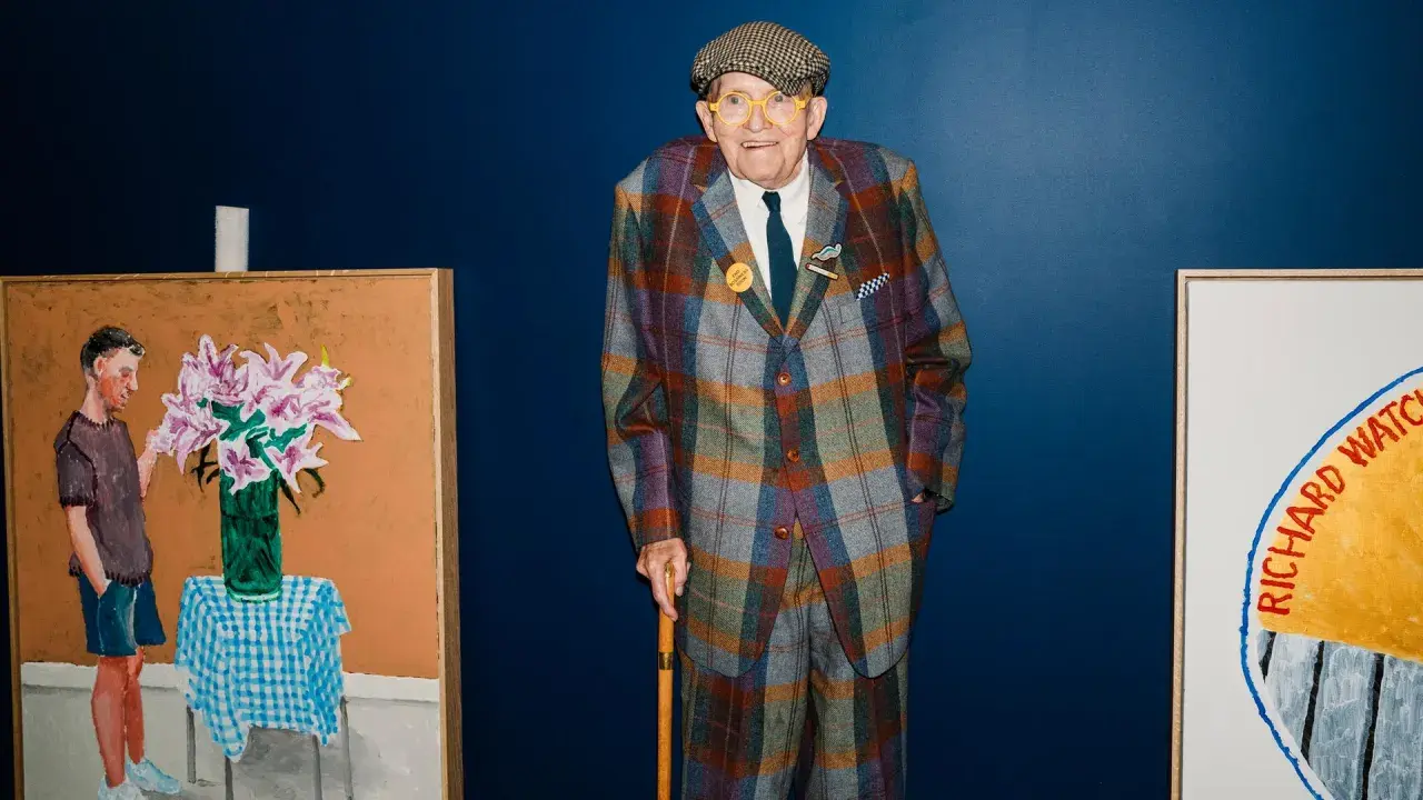 Ein Mann im karierten Anzug, inspiriert von David Hockney Pop Art, steht neben seinen lebendigen Gemälden.