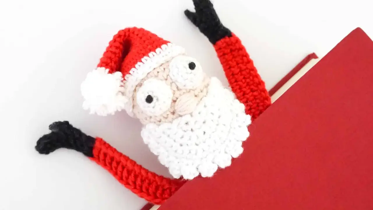 Marcador de libro amigurumi de Papá Noel, con brazos rojos y gorro festivo, asomando de un libro rojo.