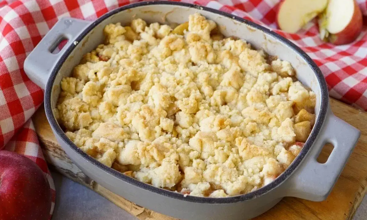 Ein goldbrauner Apfel Crumble in einer Auflaufform, bereit zum Genießen. Daneben liegen frische Äpfel auf kariertem Tuch.
