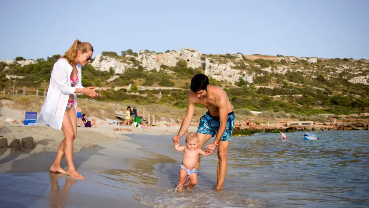 Familia disfrutando de un día en la playa en Menorca con niños. El padre ayuda a su bebé a dar sus primeros pasos en el agua, mientras la madre observa sonriente.