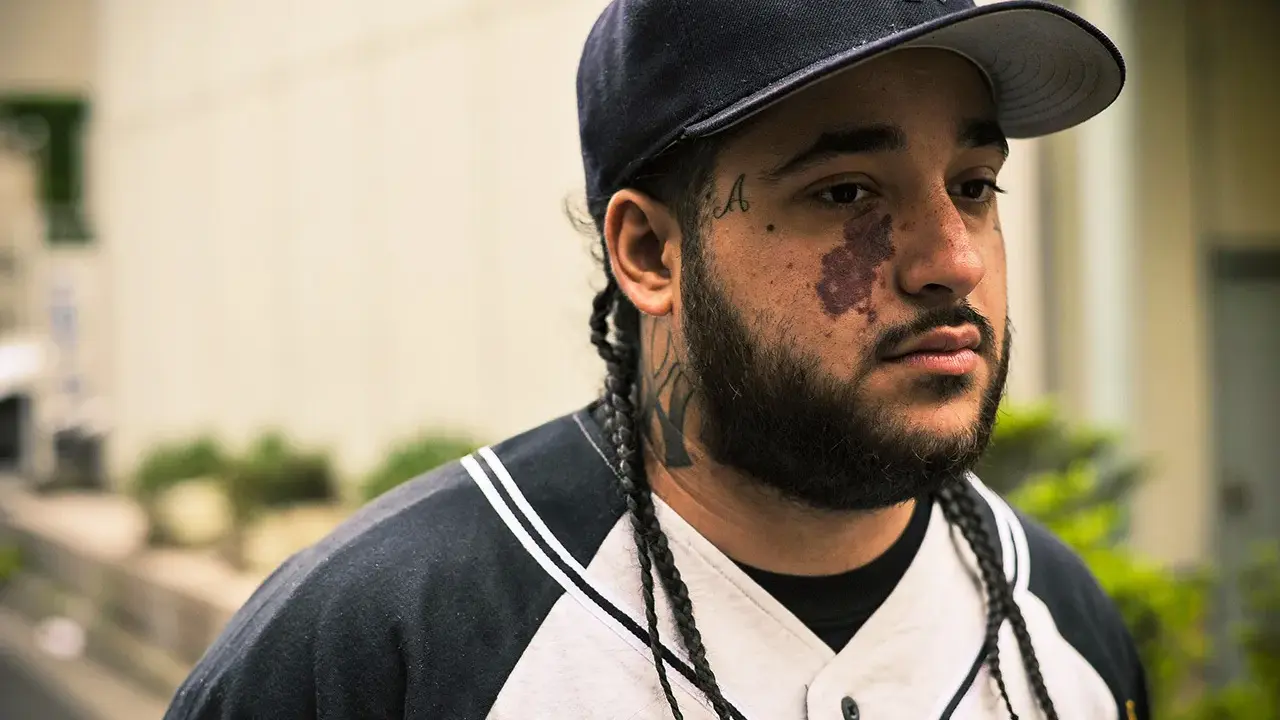 Mężczyzna w czapce z daszkiem i w bluzie z kapturem, z tatuażami na twarzy i szyi, przypominający a$ap yams.
