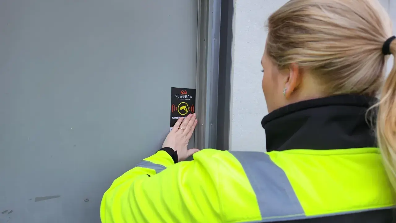 En person med reflexväst fäster en skylt med texten "Kameraövervakning" på en dörr, vilket indikerar ett larm på en byggarbetsplats.