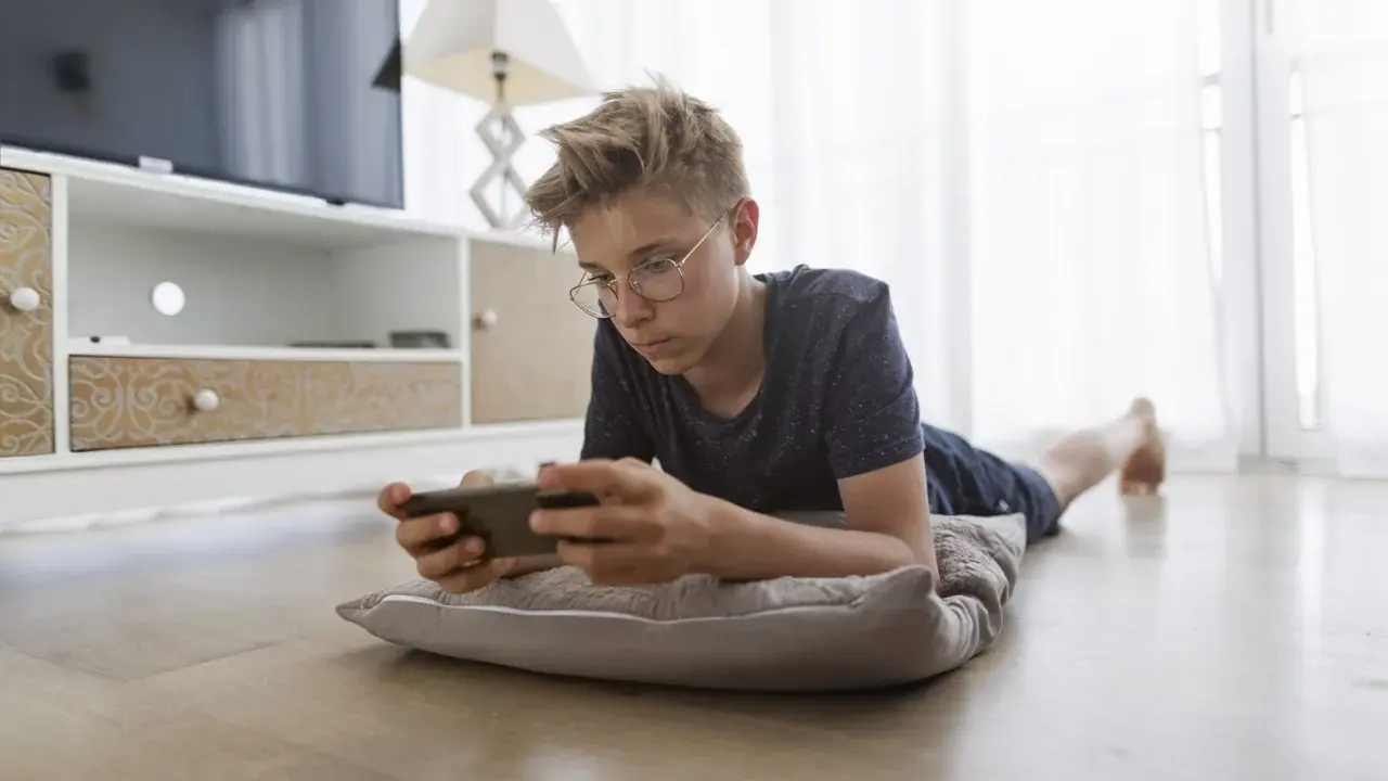 Un adolescent, absorbé par son téléphone, illustre la dépendance aux écrans. Il est allongé sur un coussin, le regard fixé sur l'appareil.
