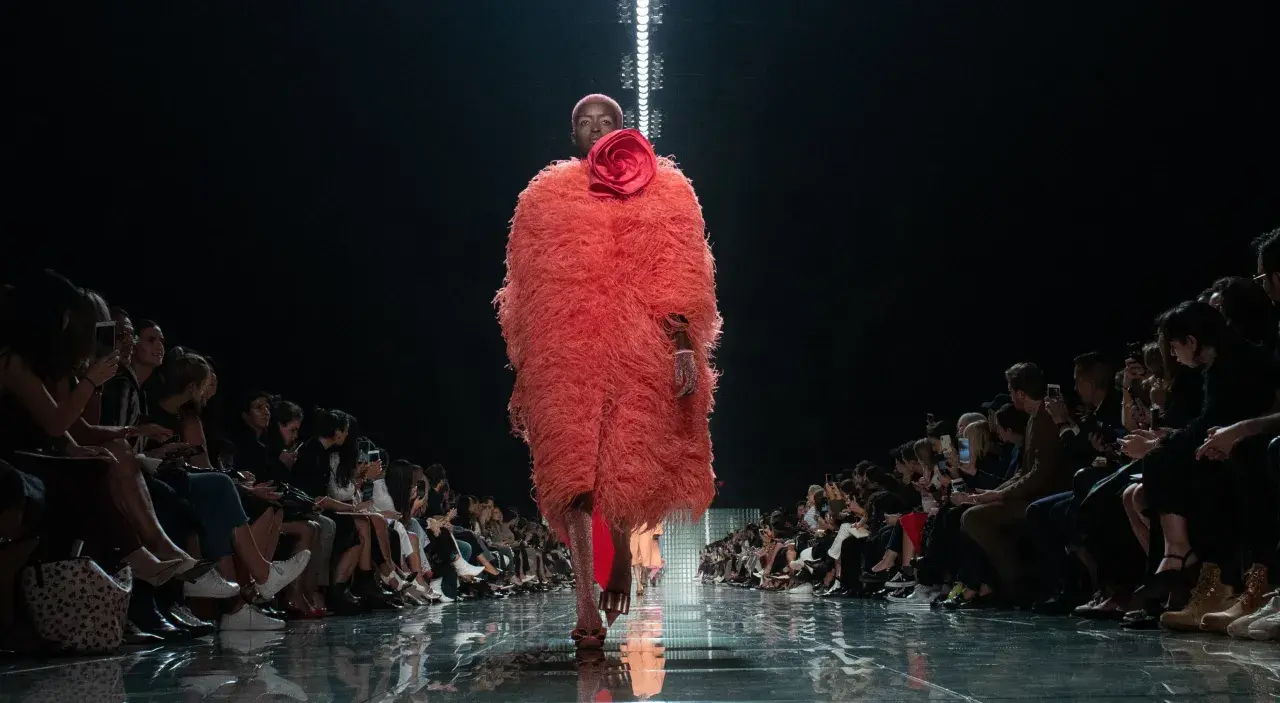Modella con cappotto vaporoso color corallo e fiore rosso, sfila su una passerella lucida. Il pubblico ammira gli abbinamenti audaci.