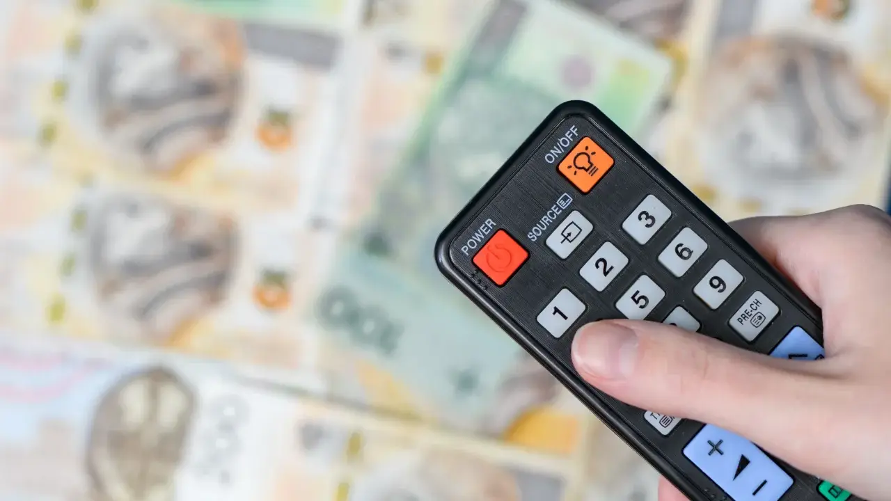 Dłoń trzyma pilota na tle polskich banknotów. Czy abonament RTV i zameldowanie mają związek z kosztami?