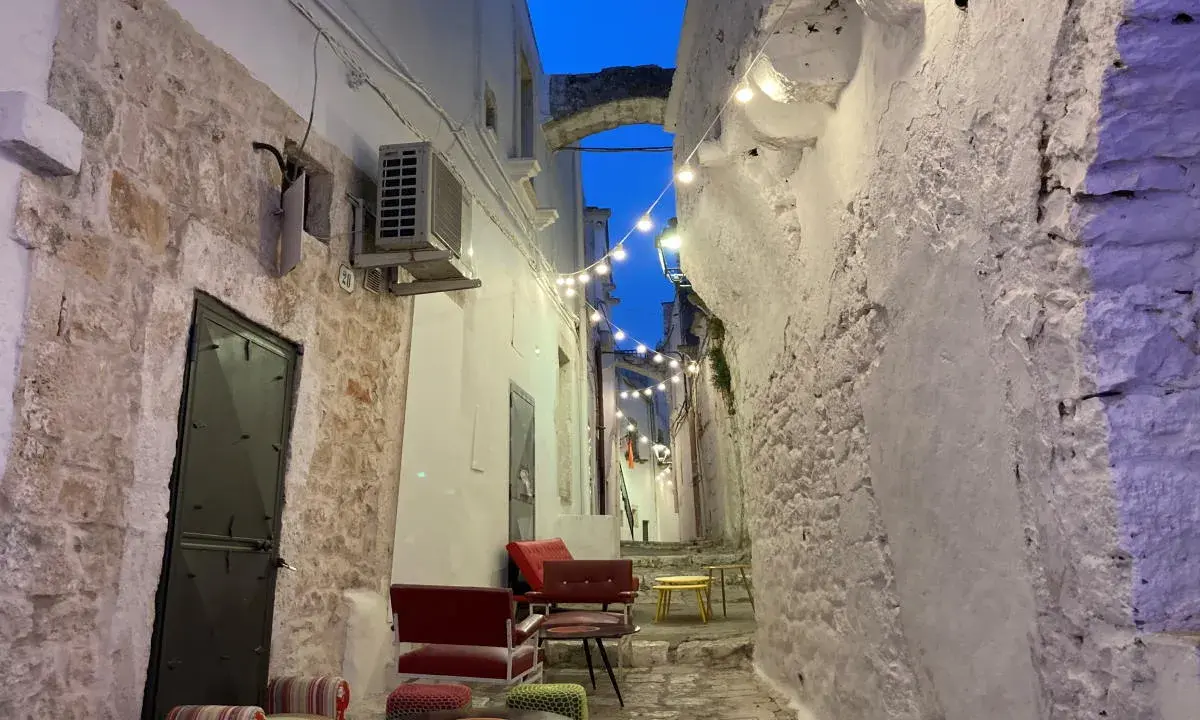 Un vicolo suggestivo nel Val d'Itria, illuminato da lucine, con sedute colorate e muri in pietra bianca.