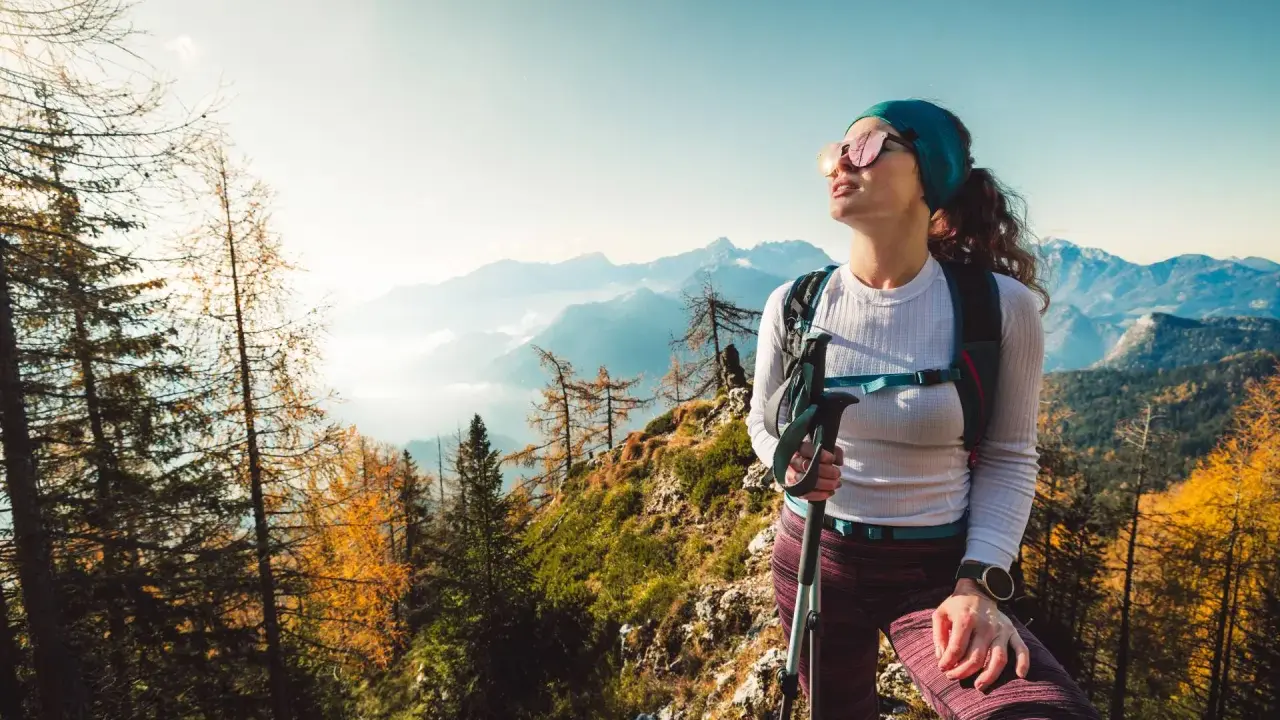 Kobieta w okularach przeciwsłonecznych i sportowym stroju, z kijkami trekkingowymi, podziwia jesienny krajobraz górski. Idealny przykład, jak się ubrać w góry.