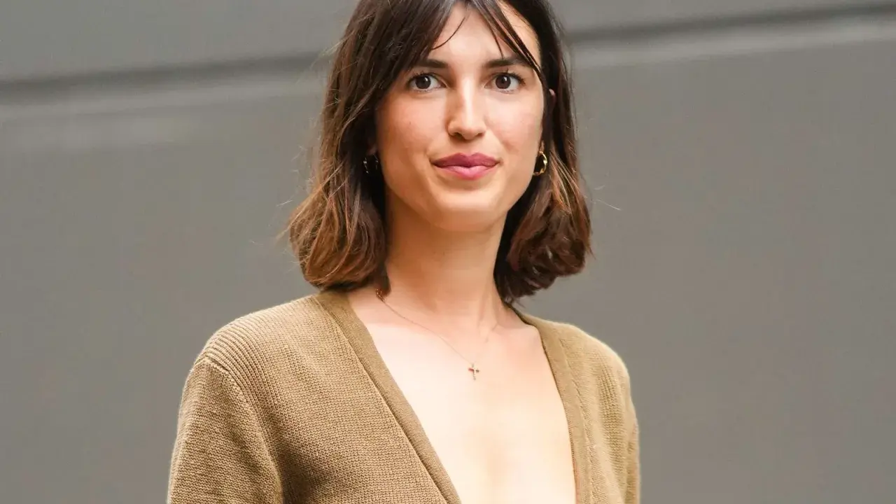 Mujer con cabello castaño ondulado, en transición de pelo corto a largo, luce un suéter marrón y un collar con cruz.