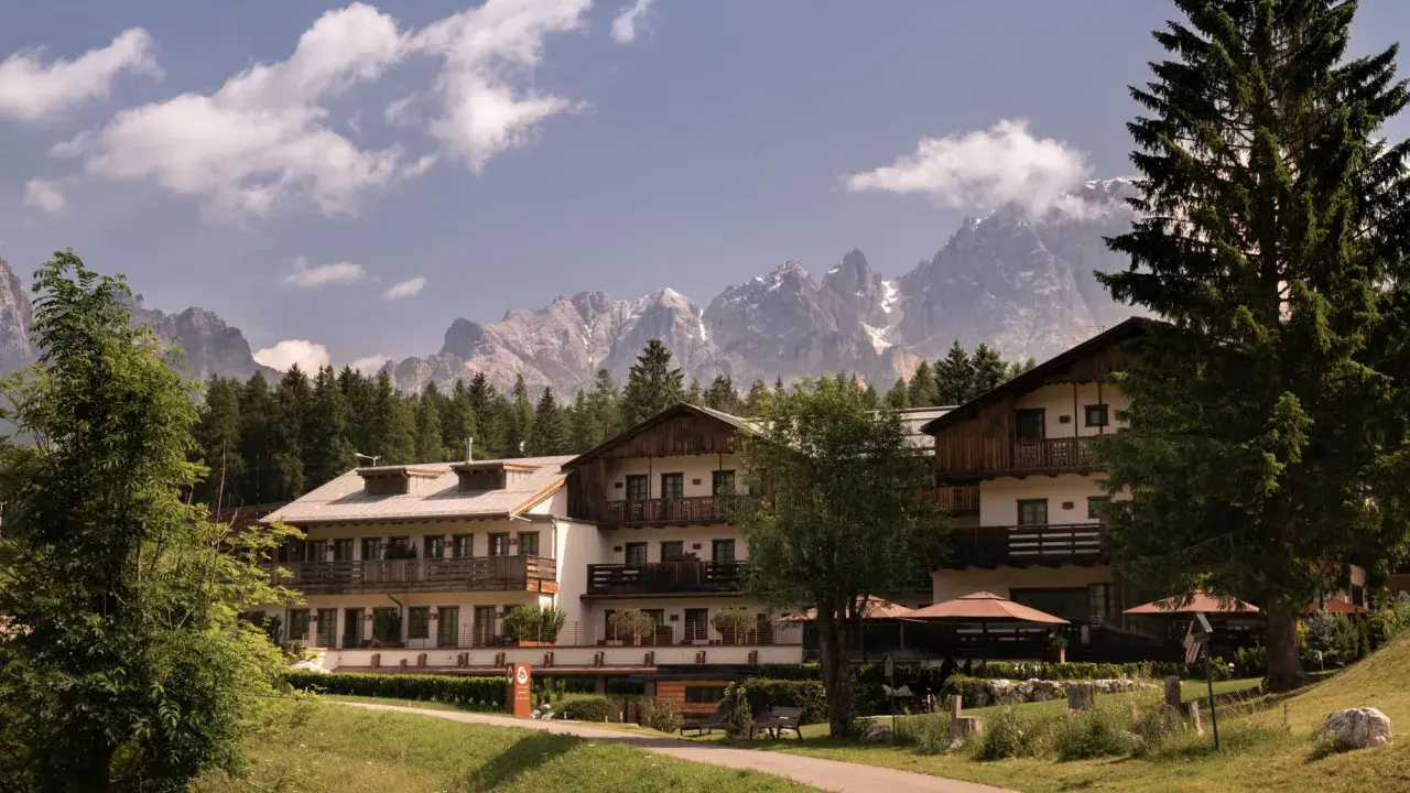 Un hotel alpino immerso nel verde, con imponenti vette innevate sullo sfondo, un perfetto scenario da montagna d'estate.