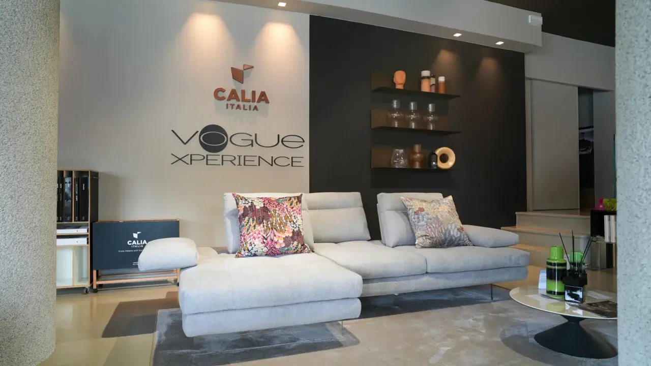 Divano componibile grigio chiaro con cuscini decorativi, logo Calia Italia e "Vogue Xperience" sulla parete.