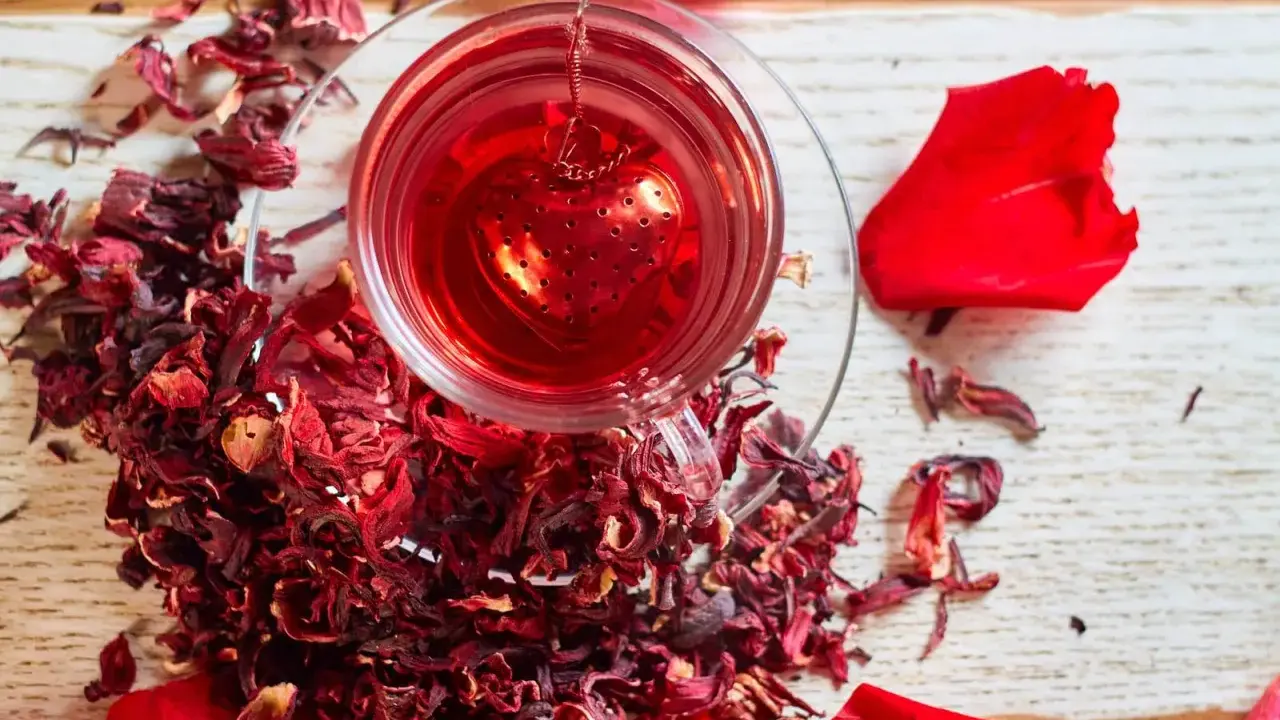 Czerwona, aromatyczna herbata z hibiskusem w szklance z zaparzaczem w kształcie serca, otoczona suszonymi kwiatami i płatkami róż.