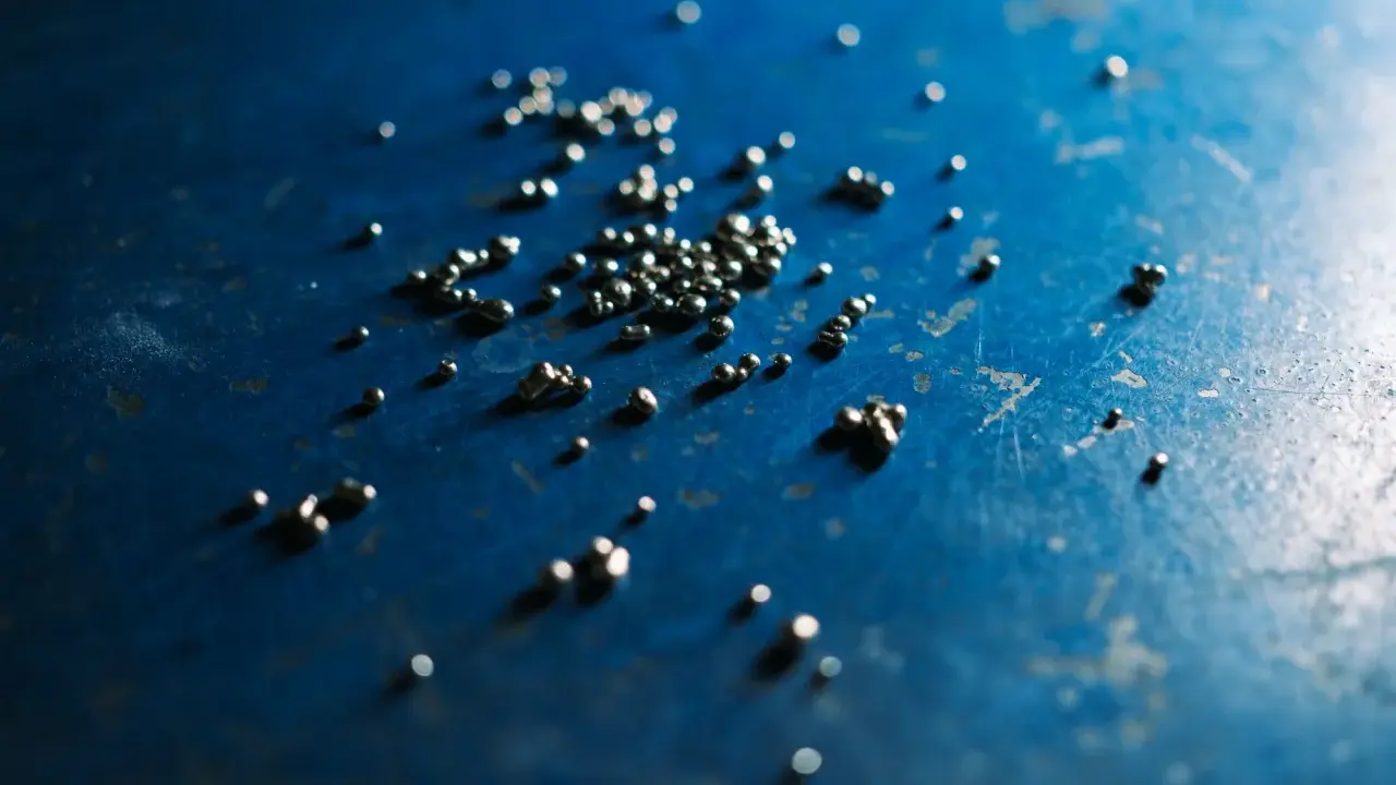 Piccole perle di argento riciclato sparse su una superficie blu scuro, con riflessi luminosi.