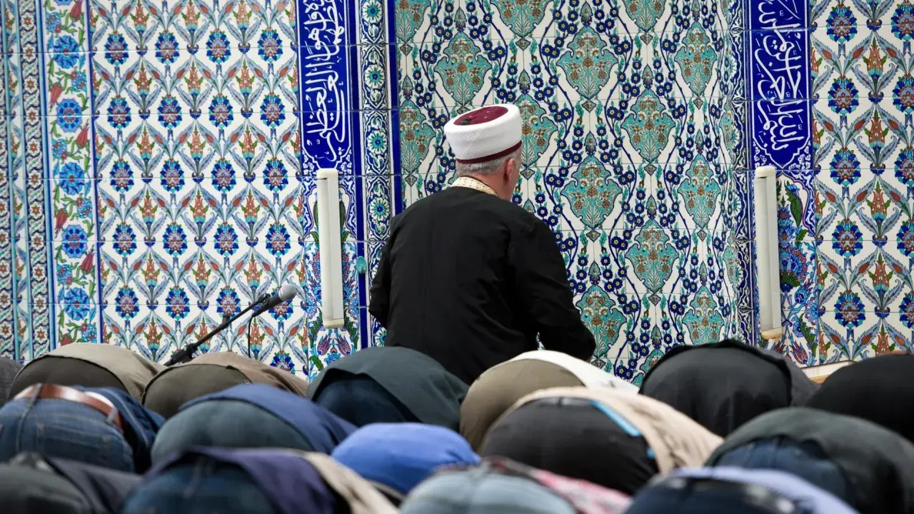 Männer in einer Moschee, die sich im Gebet verneigen. Der Imam trägt einen weißen und roten Hut. Die Fliesenwand ist mit islamischer Kalligrafie und Mustern verziert. Dies ist ein Ort, an dem der größte Sünde im Islam, Shirk, vermieden wird.