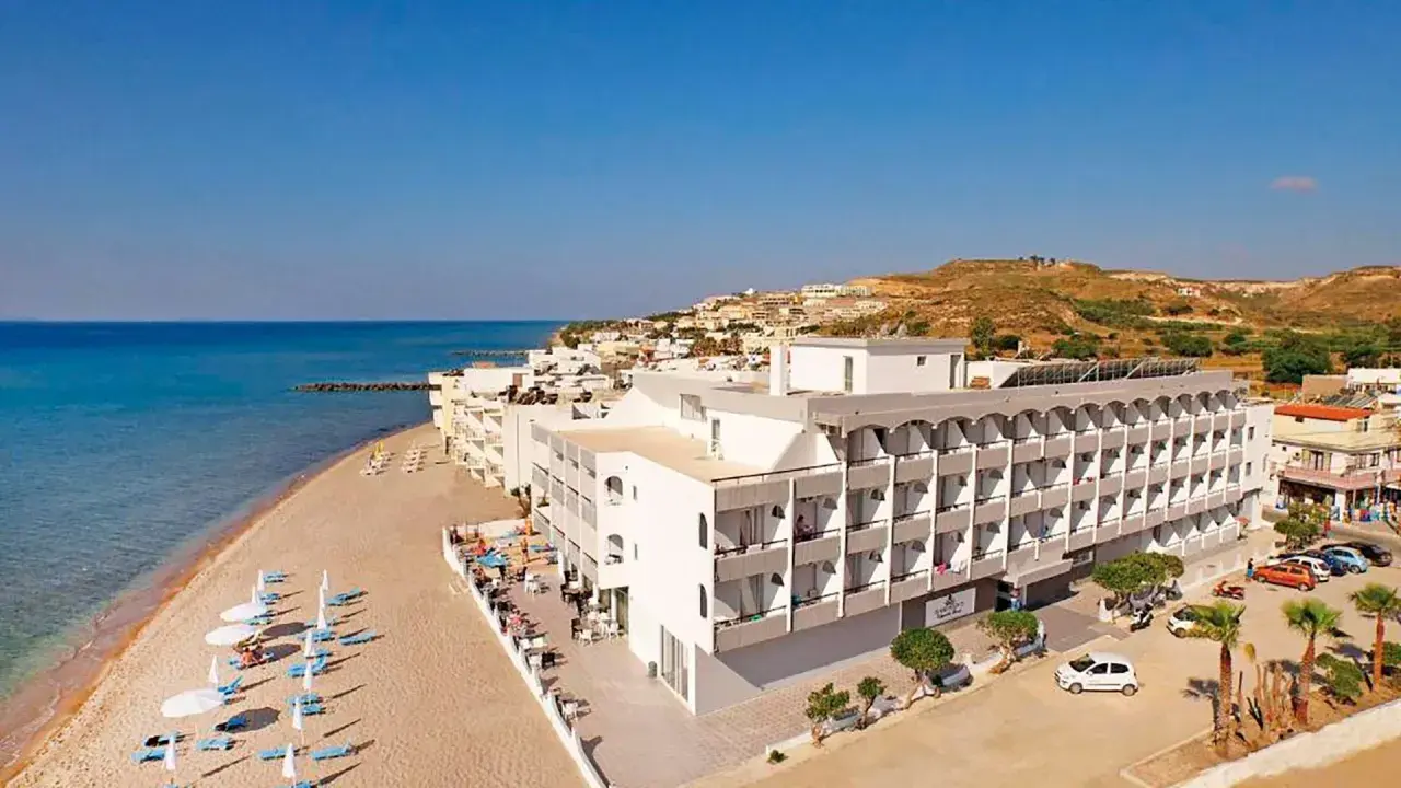 Wypoczynek na plaży w hotelu Island Maya Resort. Biały budynek hotelowy z balkonami, obok piaszczysta plaża z leżakami i parasolami, morze i wzgórza w tle.